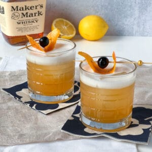 two glasses of whiskey on cocktail napkins with lemons and producers marking the whiskey in the background.