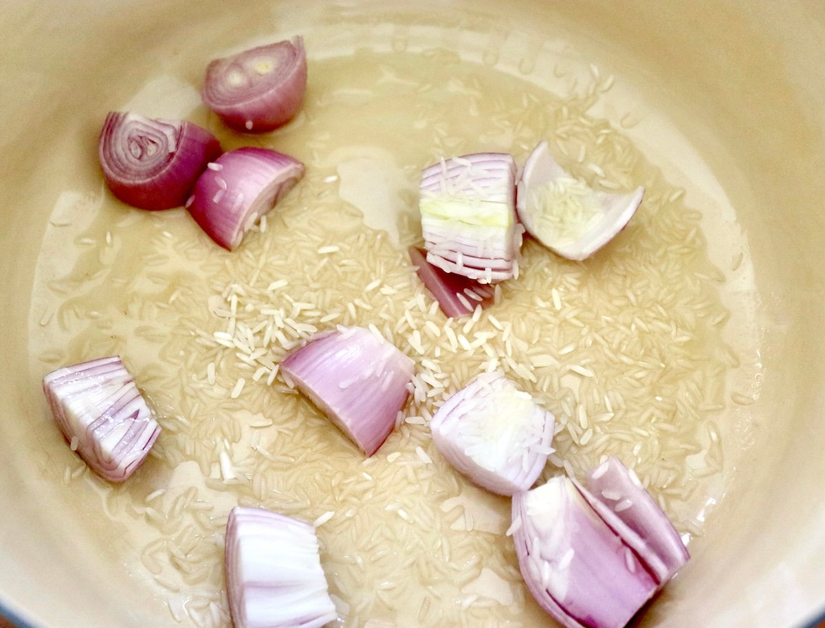 At the bottom of the dutch oven are halved shallots, sprinkled with raw rice and oil.