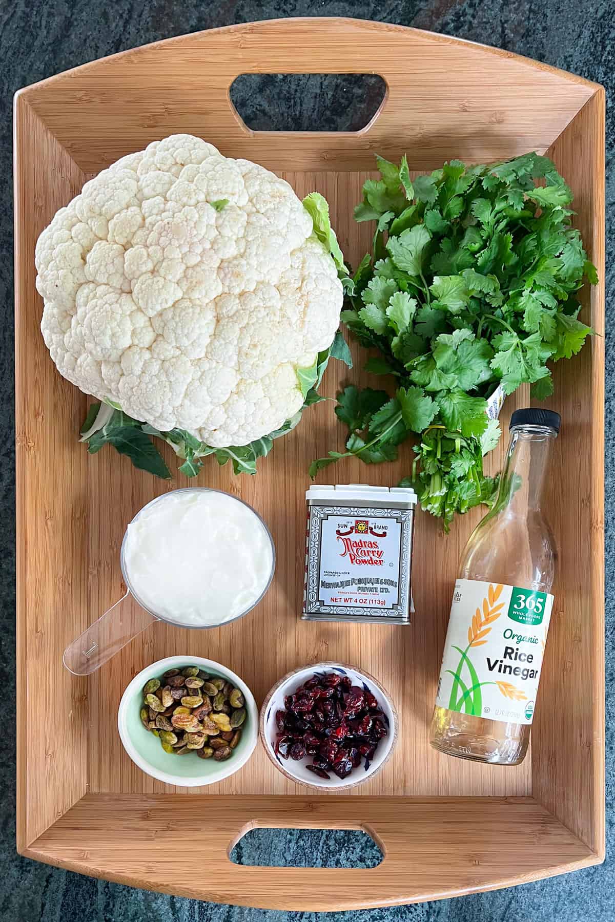 Ingredients for curried fried cauliflower with yogurt and dried cranberries, placed on a wooden tray: a head of cauliflower, a bunch of cilantro, a container of Madras curry powder, a bowl of yogurt, a bottle of rice vinegar, small bowls of pistachios and dried cranberries.