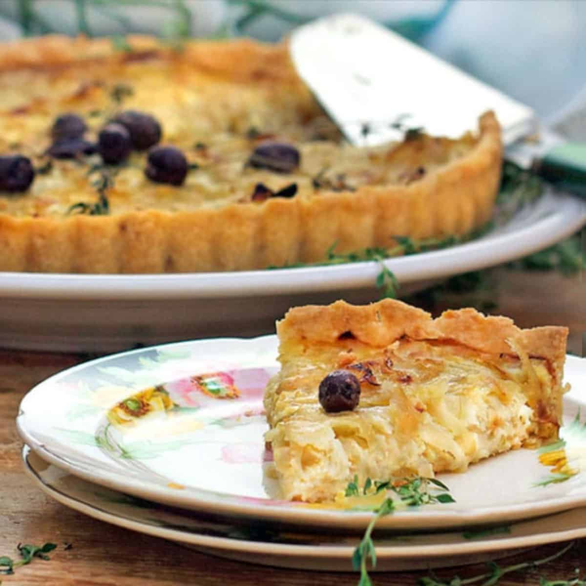 A slice of french onion tart on a fancy china appetizer plate with the rest of the tart in the background.