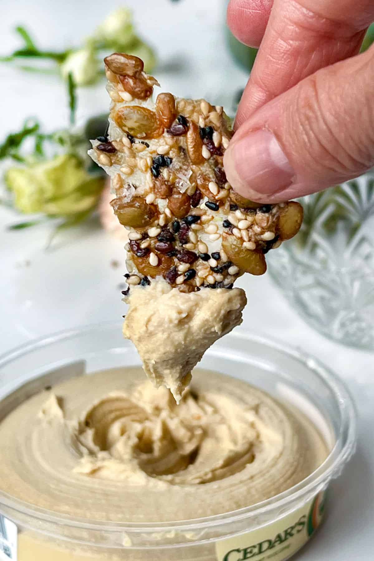 fingers holding a thin seed cracker and pulling it up out of a tub of hummus after dipping the tip.