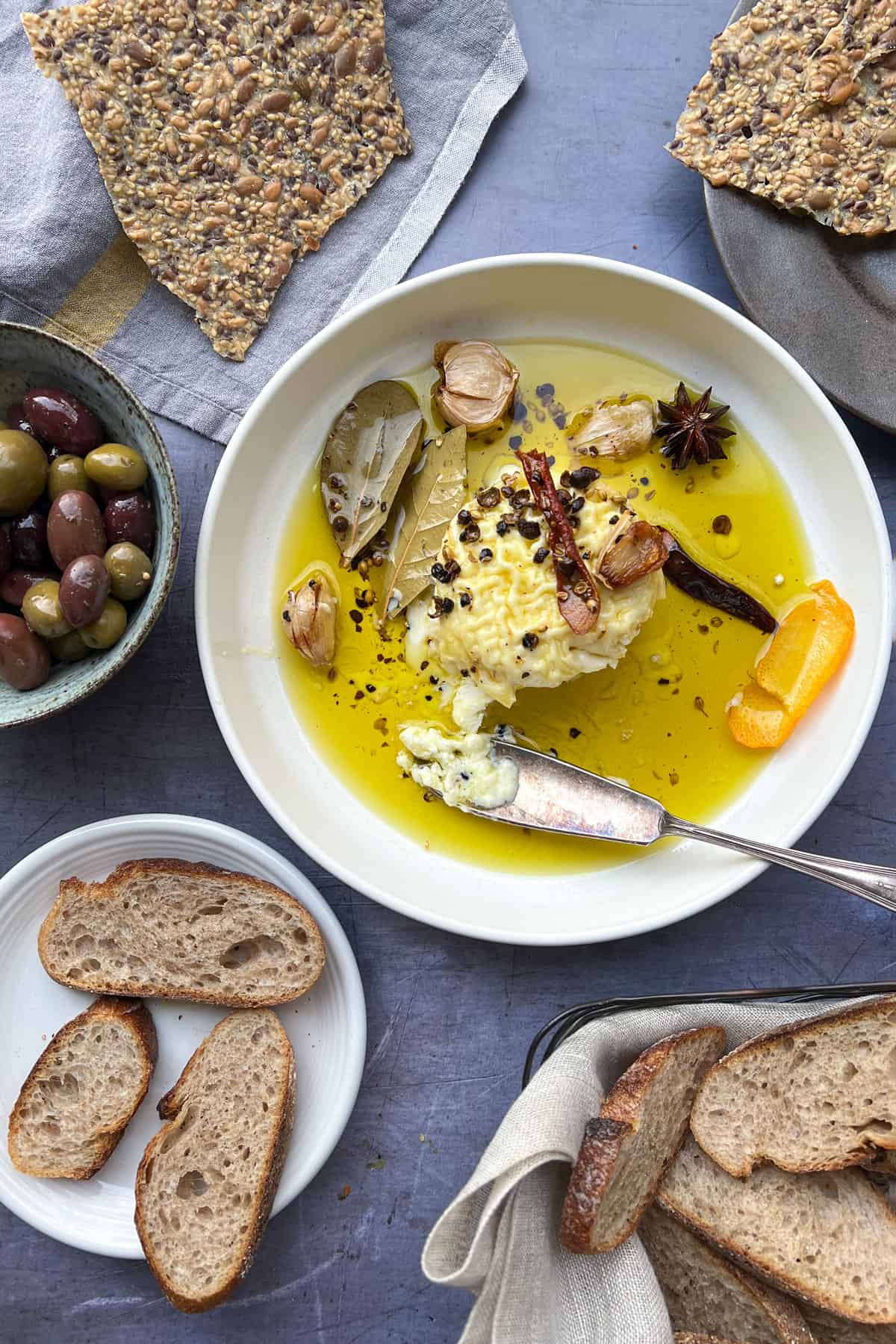 rimmed serving dish with a wheel of oil and spice covered soft cheese, surrounded by a basket of bread, a bowl of olives and some seeded crackers.