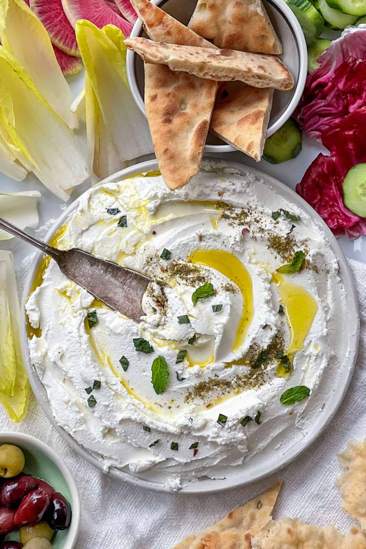 labneh dip on a plate with olive oil and mint and a knife surrounded by vegetables and pita