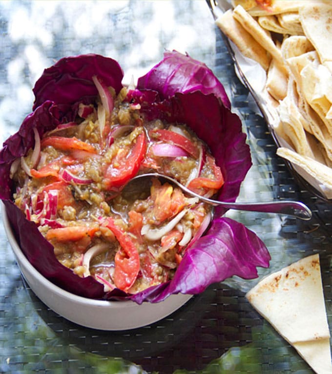 Serving bowl lined with red cabbage leaves and filled with rustic roasted eggplant dip with slices of fresh tomato and onion.