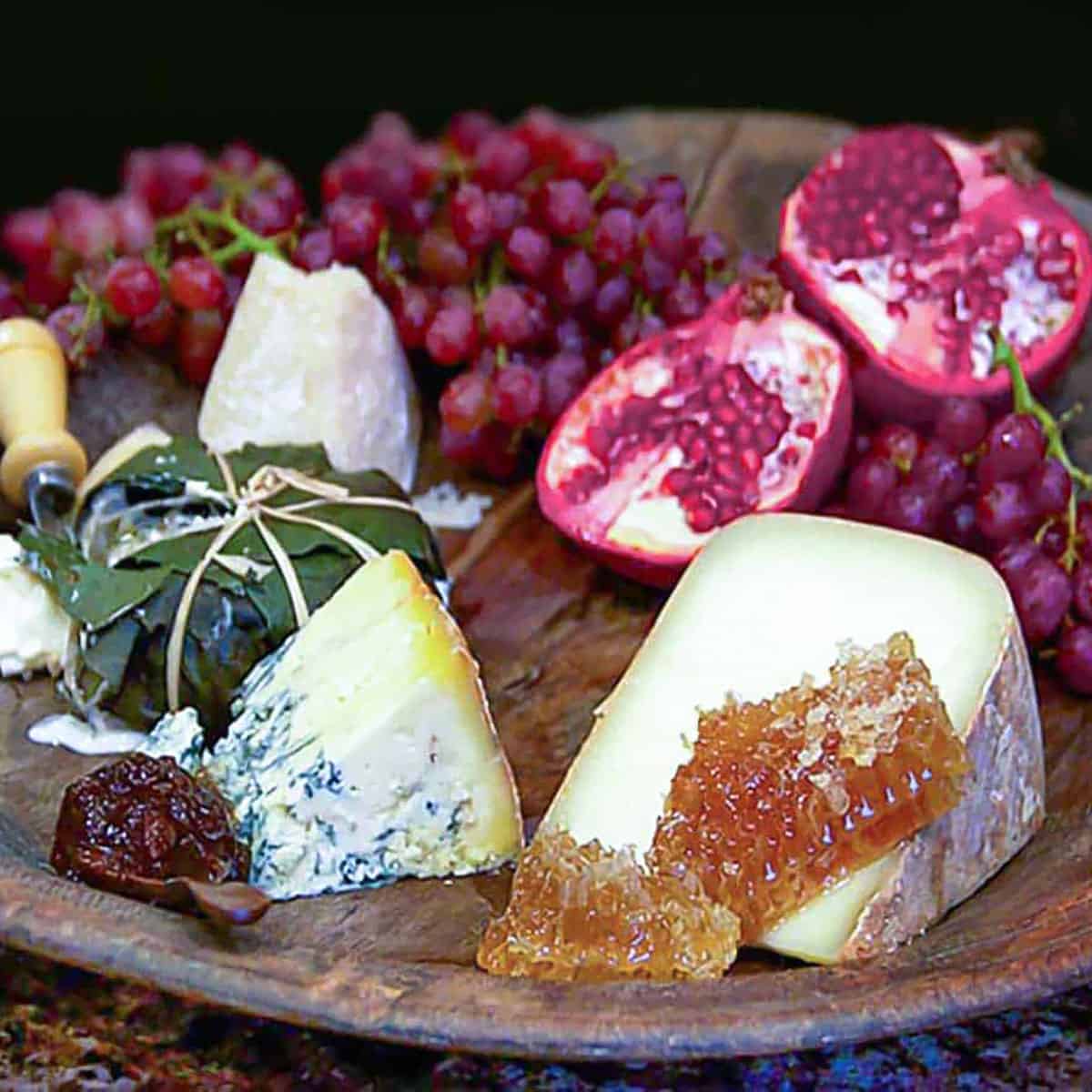 round wooden board topped with several different cheese, a bunch of grapes, a split pomegranate and a hunk of honeycomb