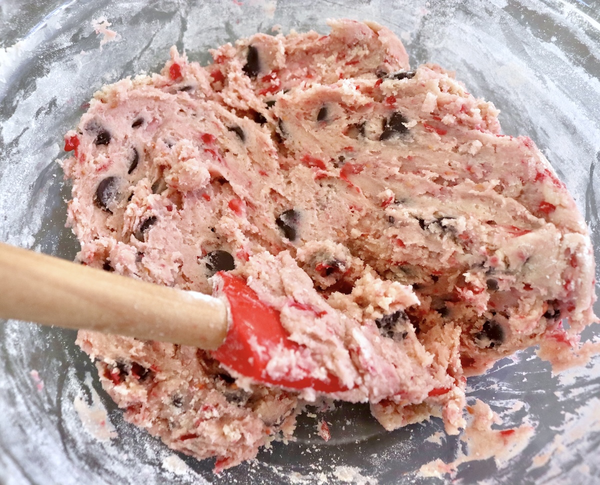 Raspberry chocolate chip cookie dough in a glass bowl with a red, wooden spatula.