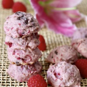 A stack of 4 raspberry chocolate chip cookies with a pink lily flower behind them, with a few bright red raspberries around them.