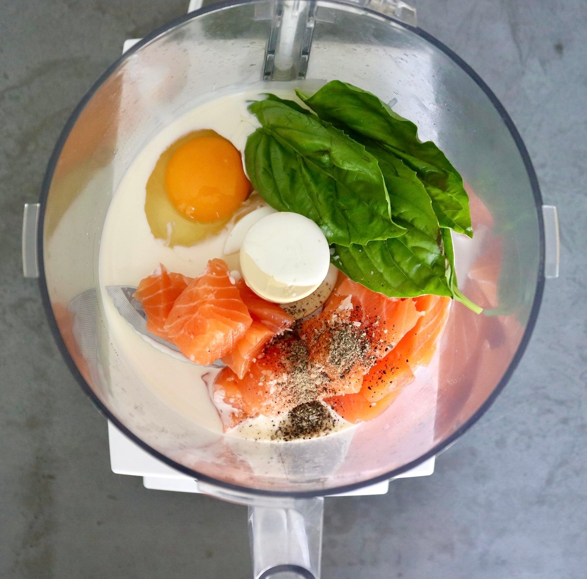 Food processor bowl with raw salmon chunks, heavy cream, egg, basil leaves, salt and pepper.