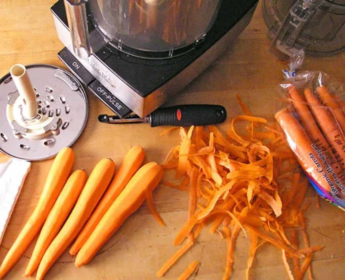 a bunch of peeled carrots on a wooden table next to a food processor and a shredding blade for a processor.