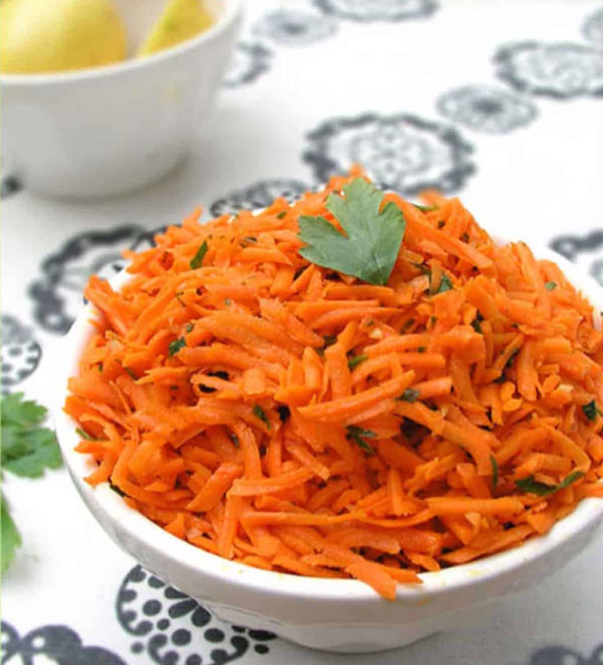 Shredded carrot salad in a bowl with a sprig of parsley. The lemon bowl in the background.