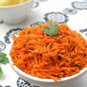 Shredded carrot salad in a bowl with a sprig of parsley. The lemon bowl in the background.