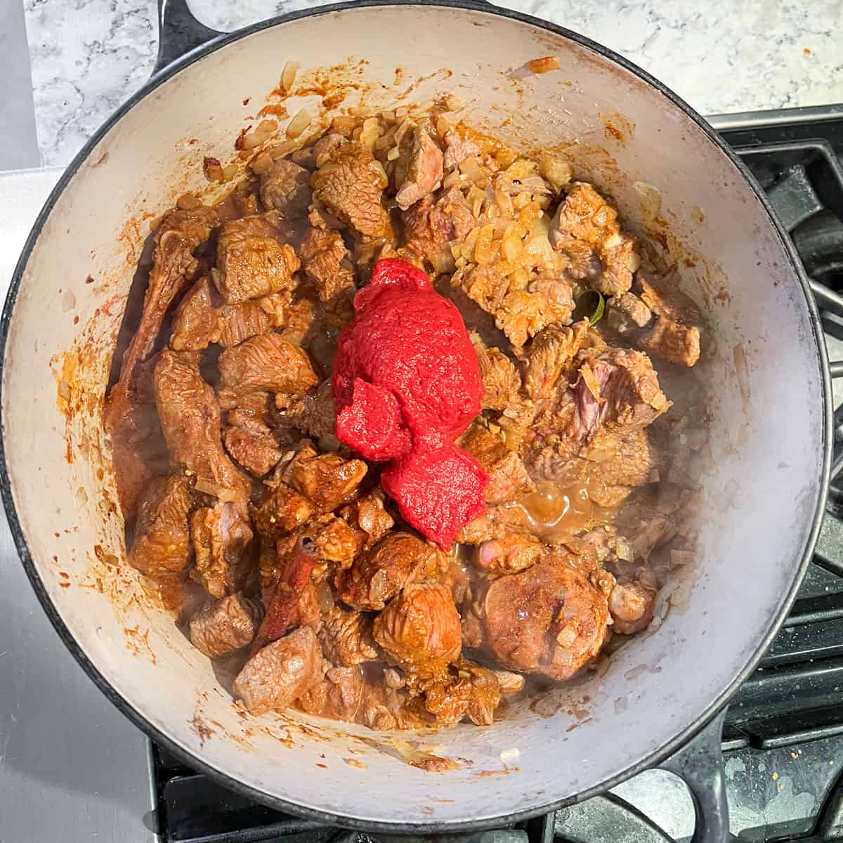 Dutch oven with cubes of lamb coated with sauce and spices and topped with a heap of tomato paste.