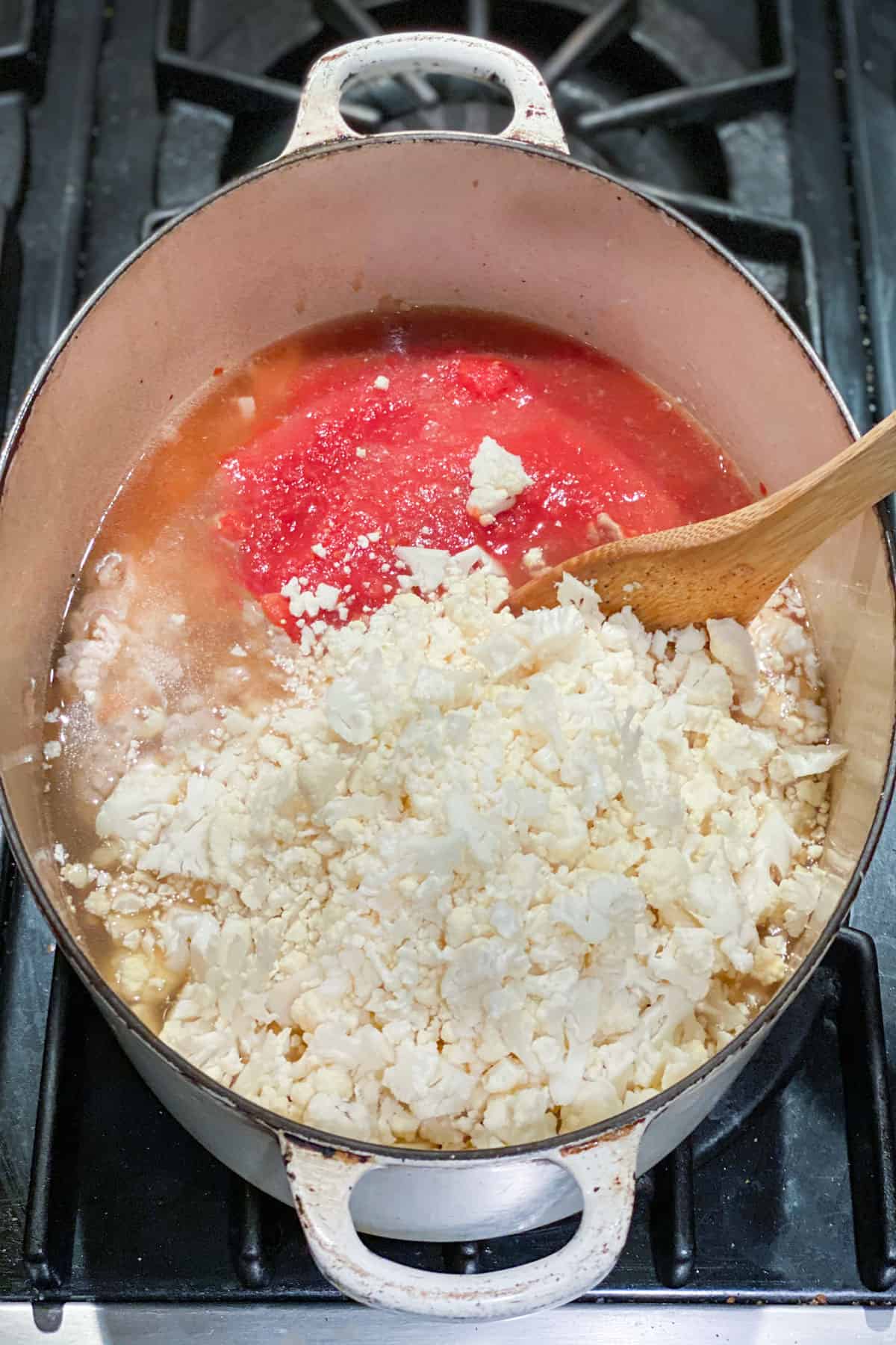 Chopped cauliflower and tomato puree being stirred with a wooden spoon, in a dutch oven.