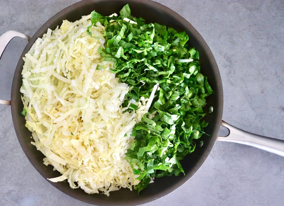 A large skillet with a pile of shredded Napa cabbage and sliced ​​Gai Lan on top of the sautéed onions.