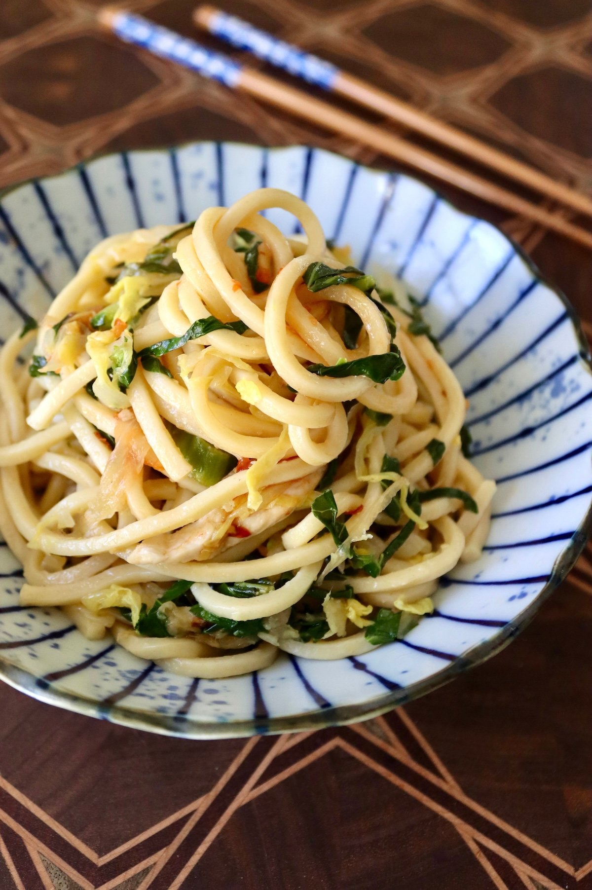 Swirled pile of Chinese Longevity Noodles with chicken and vegetables on an Asian style plate with wooden chopsticks behind it.