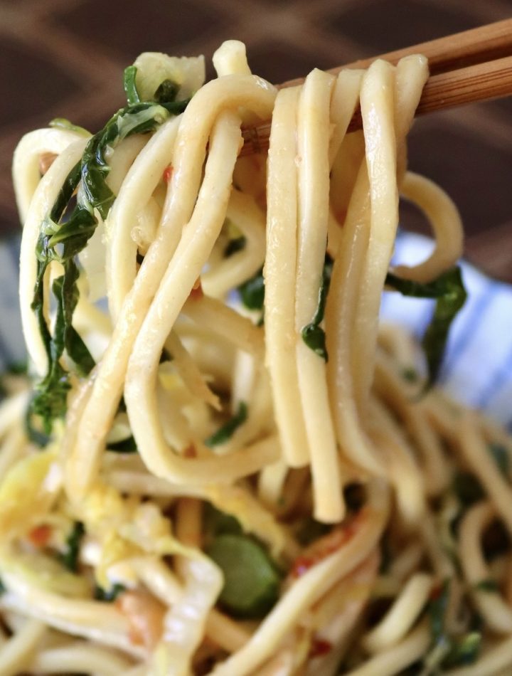 Serving of Chinese Longevity Noodles with Gai Lan on ceramic blue and white plate with a bite held up in chopsticks.