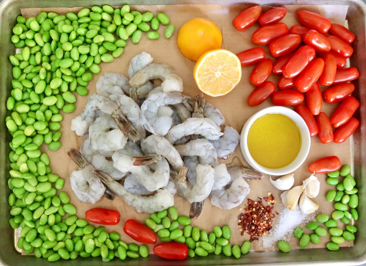 Parchment paper-lined sheet pan with fresh edamame (shelled), raw shrimp, small tomatoes, a plate of olive oil, garlic cloves, crushed red pepper and lemon - all placed separately.