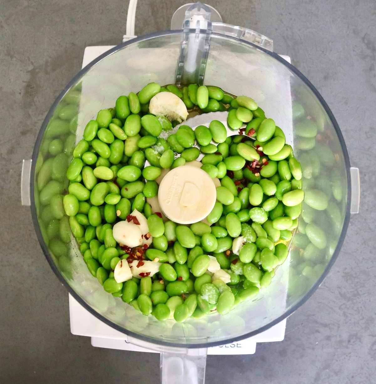 Food processor bowl with blade attachment and edamame, raw garlic, and crushed chili.