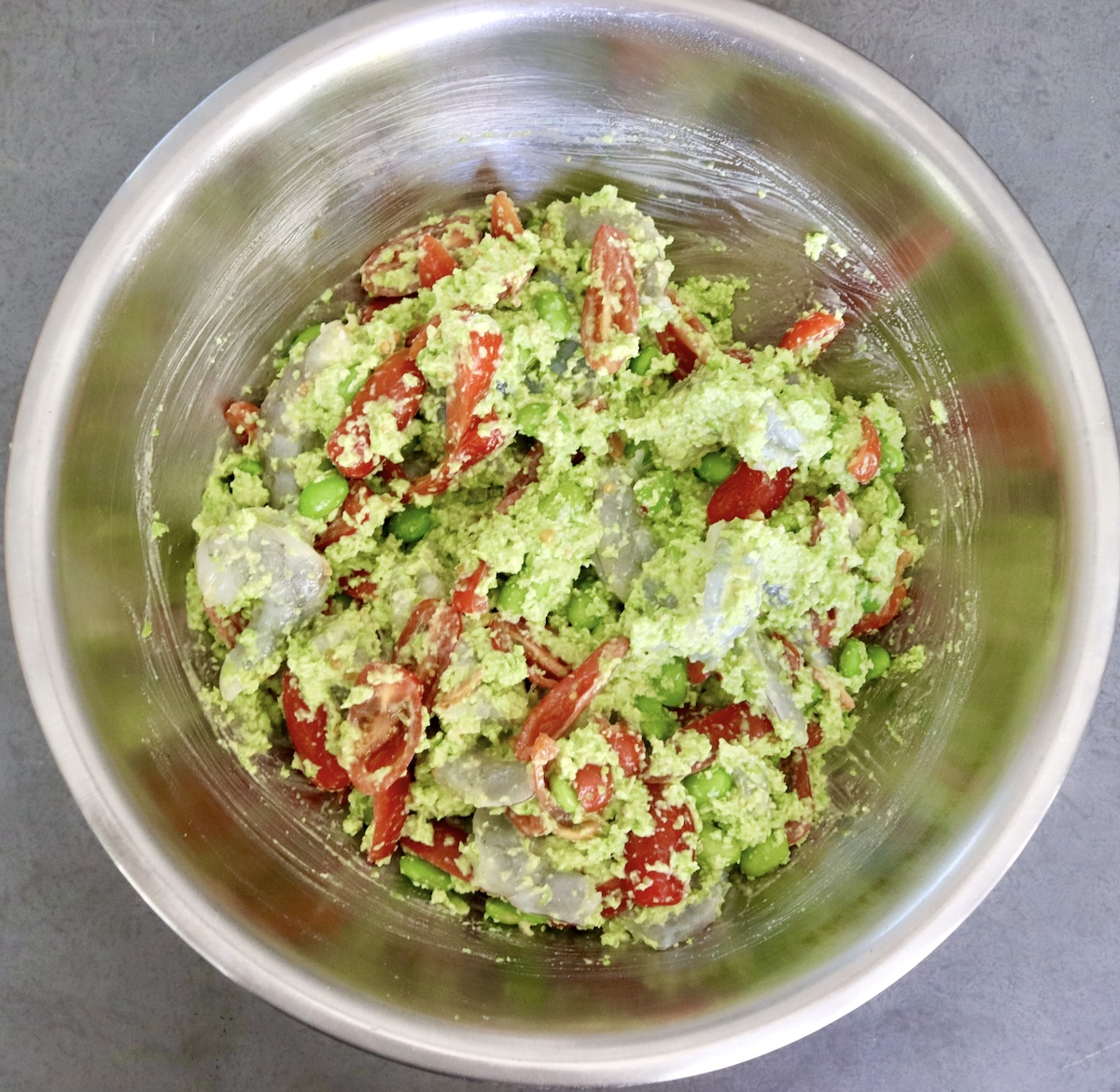 A stainless steel mixing bowl with a mixture of raw shrimp, edamame pesto and chopped tomatoes.