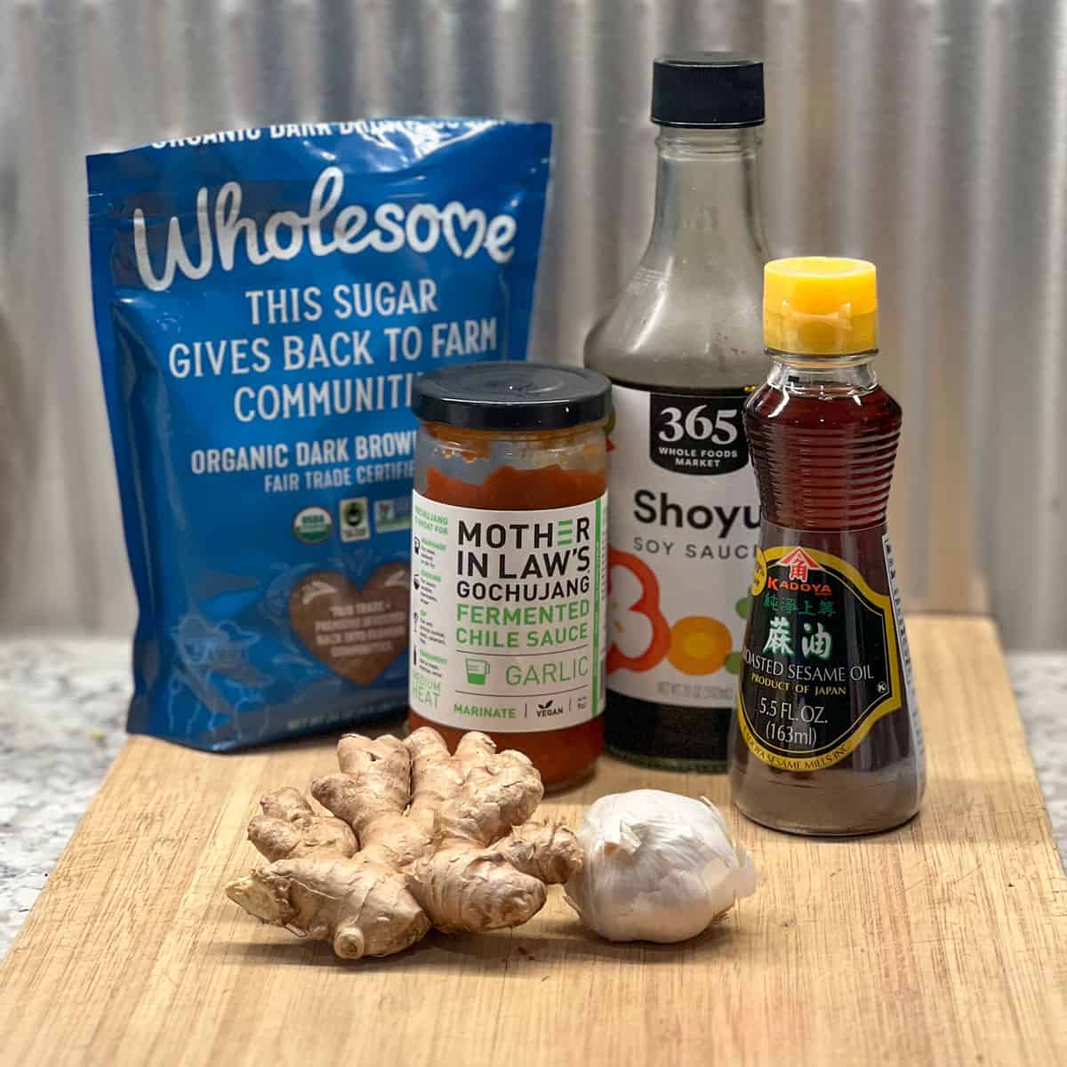 Ingredients on a wooden cutting board, a bag of brown sugar, bottles of soy sauce, gochujang, and sesame oil, a hunk of ginger root, a whole clove of garlic.