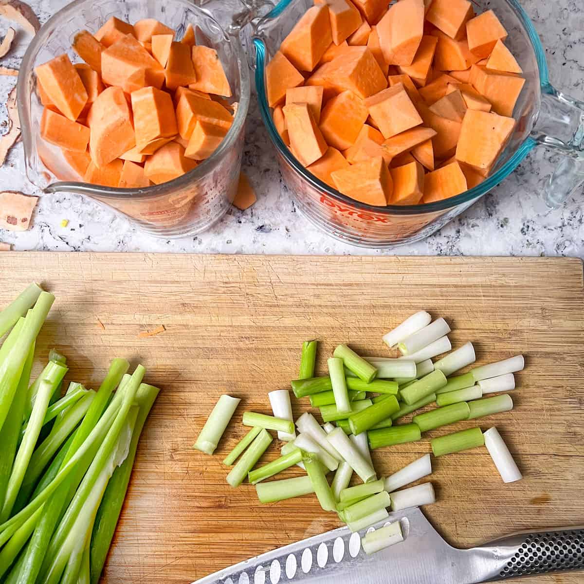 two large measuring cups filled with cubed sweet potatoes next to a cutting board with a knife and chop the scallions.