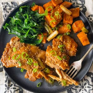 Two cooked chicken thighs topped with scallions and sesame seeds, with cubes of fried potato and cooked spinach on the plate.