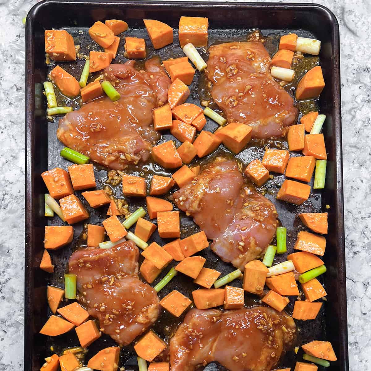 raw marinated boneless chicken thighs arranged on a sheet pan with cubed sweet potatoes and scallions.