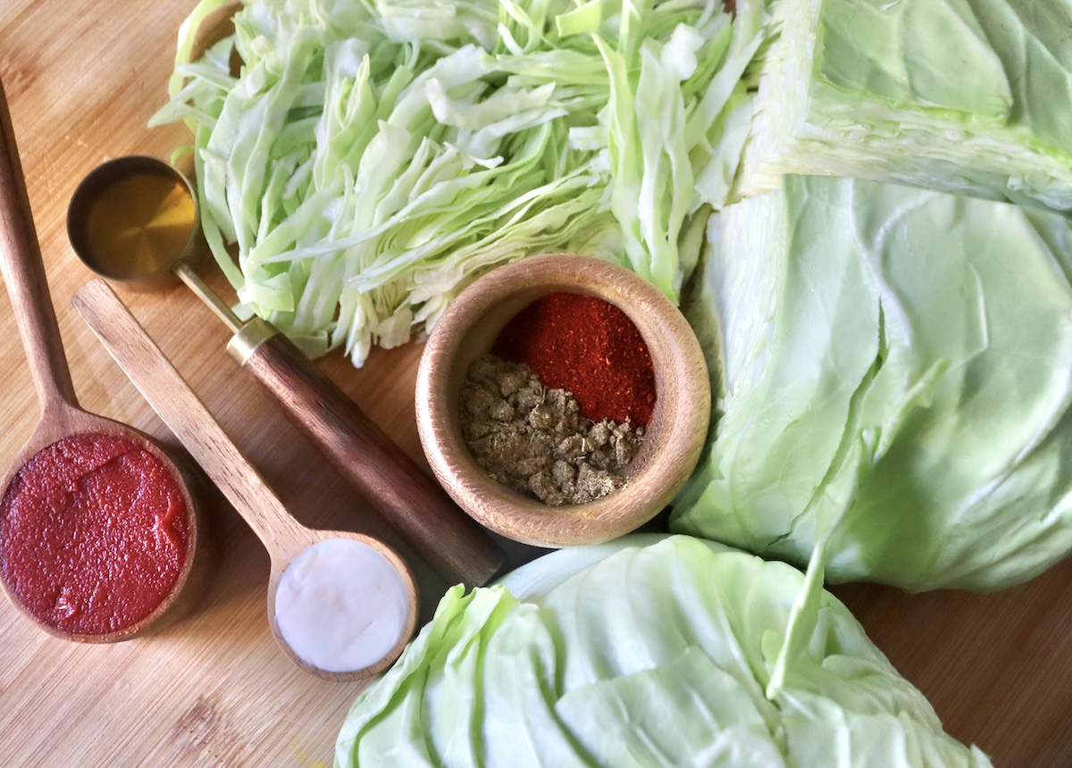 Ingredients for tomato sautéed cabbage include green cabbage, paprika, cumin, butter, tomato paste and olive oil, on a wooden cutting board.