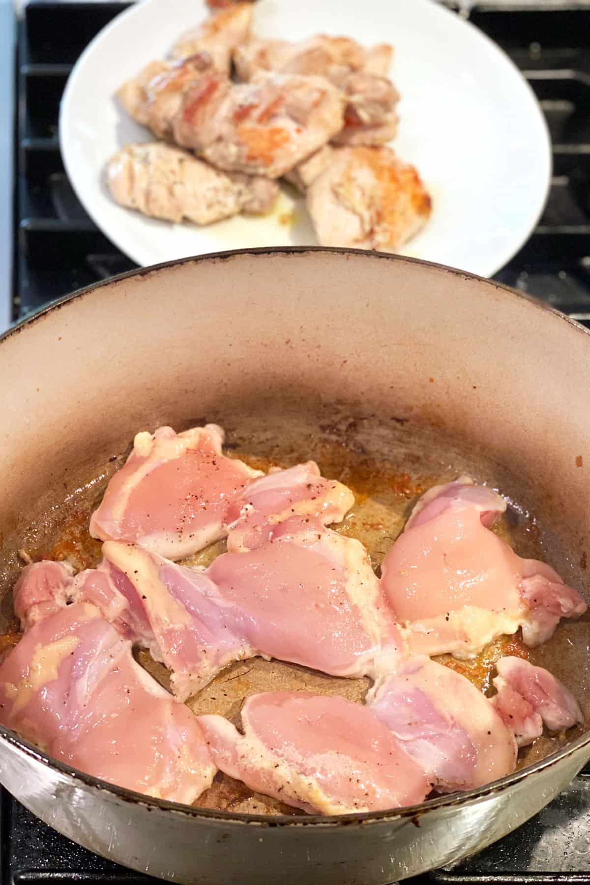 several boneless chicken thighs browning in a dutch oven with a plate in the back holding a few boneless chicken thighs.