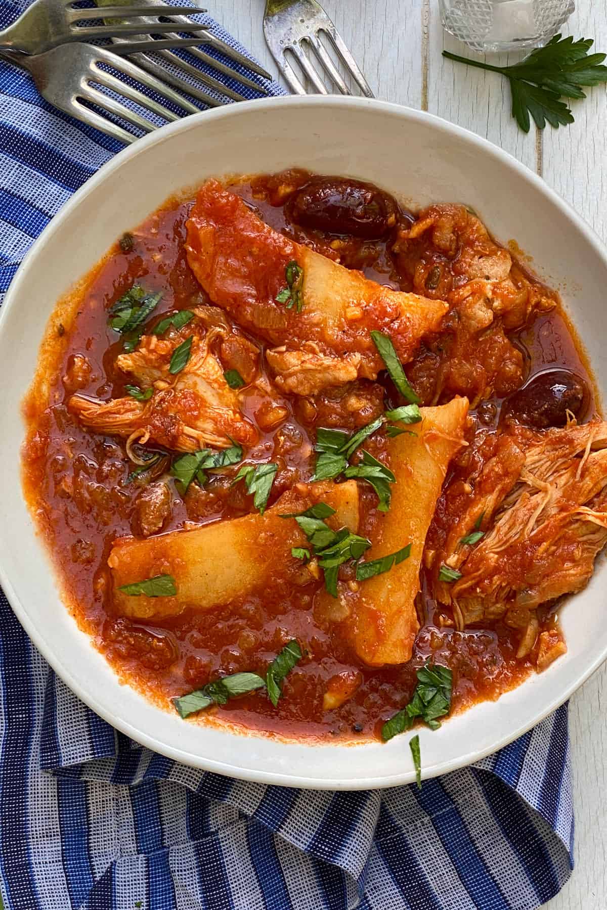a white bowl on a blue striped dish, filled with Greek chicken stew with potatoes and olives in a tomato sauce