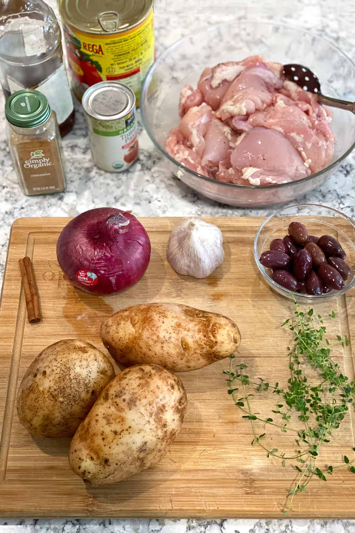 Ingredients on the granite countertop: a glass bowl of boneless chicken thighs, 3 russet potatoes, 1 red onion, a few thyme leaves, a small bowl of kalamata olives, a cinnamon stick, a head of garlic, a can of tomato paste, a can of plum tomatoes and a jar of cinnamon.