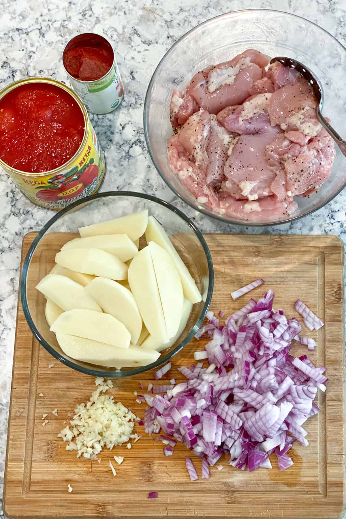 Items prepared on the countertop: Potato wedges in a glass bowl, Sliced ​​red onion in a bowl, chopped garlic on a cutting board, opened canned tomatoes, boneless chicken thighs seasoned with salt and pepper in a bowl.