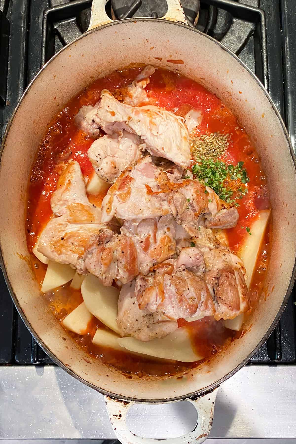 An oval dutch oven filled with browned chicken thighs, potato wedges, tomato sauce and spices.