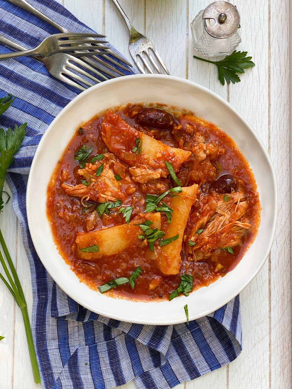 a white bowl on a blue striped dish, filled with Greek chicken stew with potatoes and olives in a tomato sauce