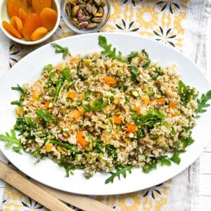 a bowl of rice and quinoa salad with a small bowl of dried apricots and a small bowl of roasted pistachios in the background.