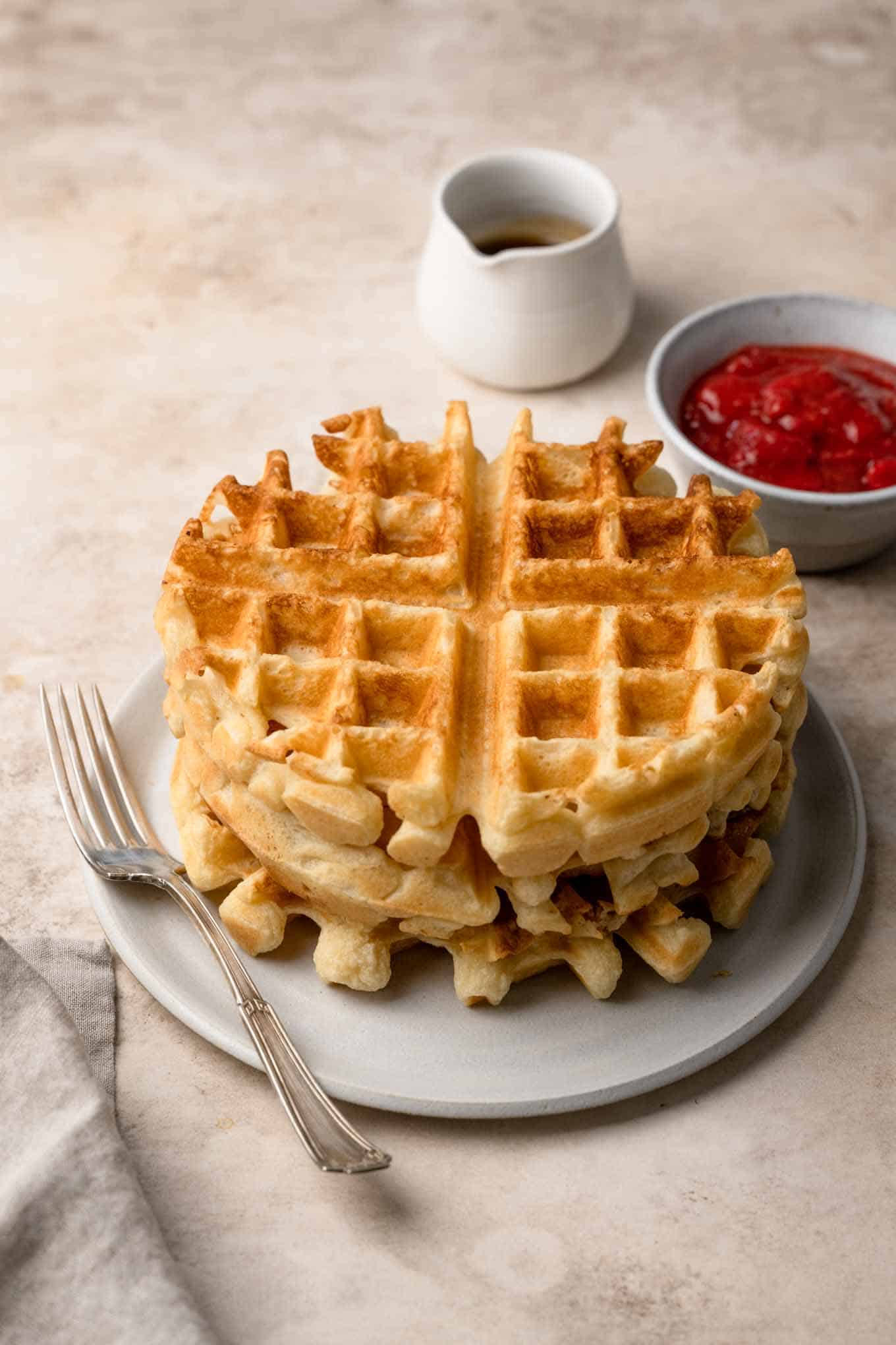 Three waffles stacked in a bowl with a jug of maple syrup and fruit compote to serve