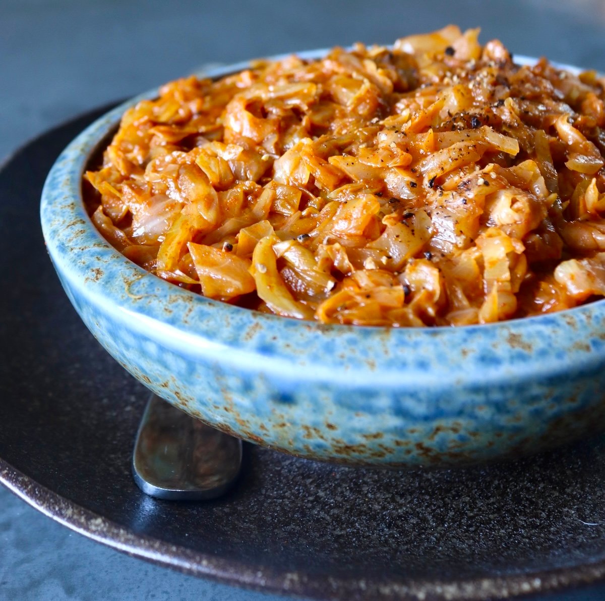 A light blue ceramic bowl full of tomatoes and cooked cabbage, on a black ceramic plate.