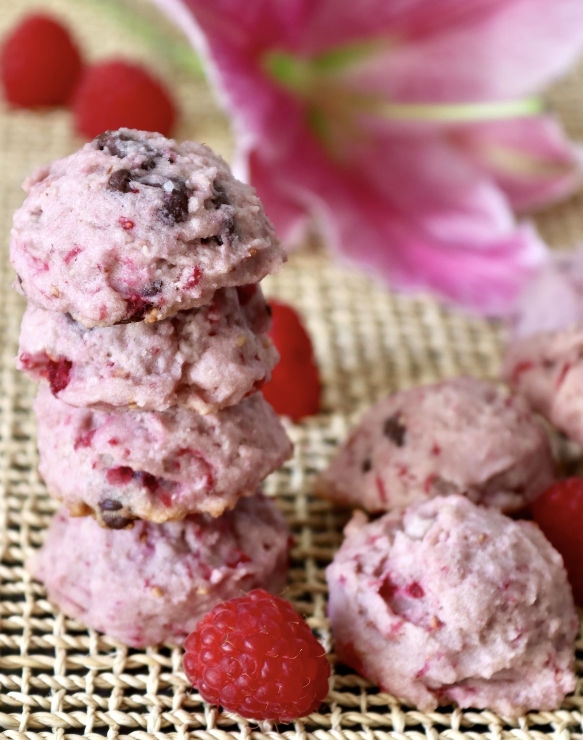 A stack of 4 raspberry chocolate chip cookies with a pink lily flower behind them, with a few bright red raspberries around them.